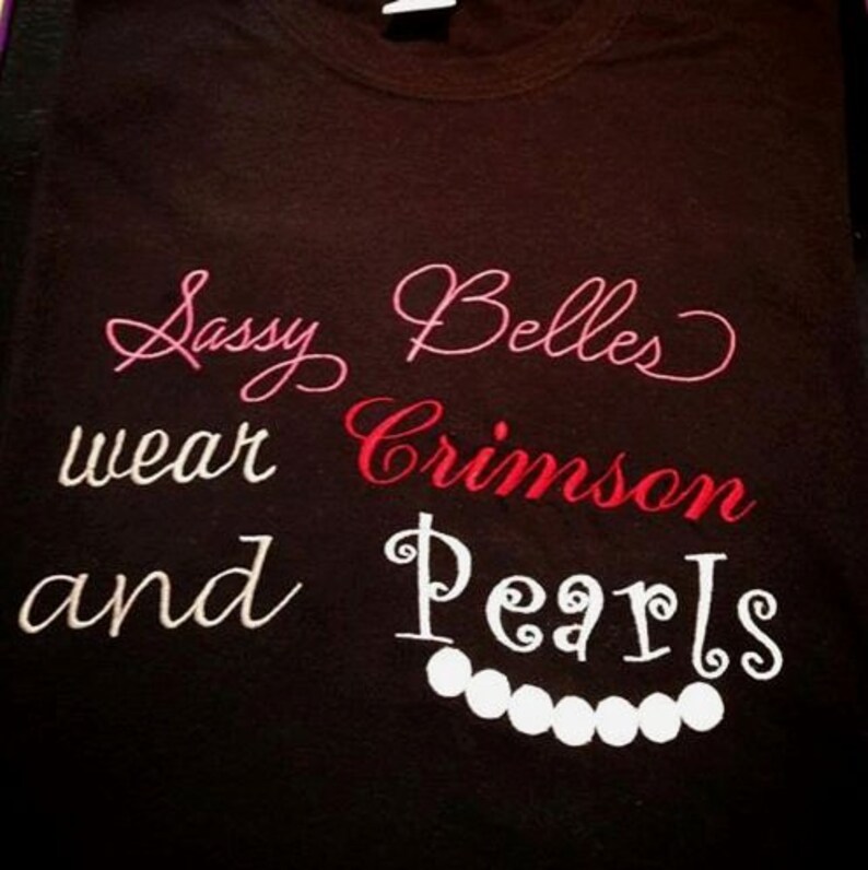 May include: Black t-shirt with white embroidery that reads "Sassy Belles wear Crimson and Pearls" with a string of white pearls below the text.