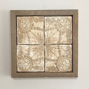May include: A framed wall decor with four square tiles featuring a beige and white floral pattern. The tiles are arranged in a grid pattern and have a raised, textured design.