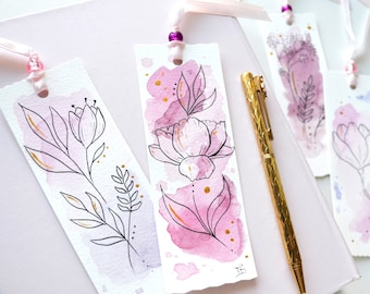 Pode incluir: Três marcadores de livros de aquarela rosa com detalhes dourados e desenhos florais de tinta preta. Os marcadores estão sobre uma superfície branca com uma caneta dourada.