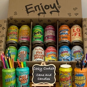 May include: A collection of colourful, miniature cans and candles with various designs and names, including "Cwtch", "Cali Pale", "Key Lime Lager", "Five Dollar Shake", "Rhubarb Custard Sour", "Electric Boogaloo", "Lo-Fi", "Peaches and Cream", "Lime", "Easy Livin", "Dutty", "Switch", and "Shake Down". The cans and candles are arranged in a box with the words "Enjoy!" and "Cosy Cwtch Cans and Candles" written on it.
