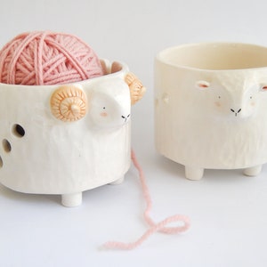 May include: Two white ceramic yarn bowls shaped like sheep. One bowl has a pink ball of yarn inside and a strand of yarn hanging out.