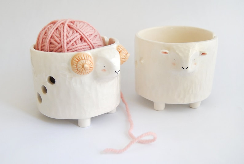 May include: Two white ceramic yarn bowls shaped like sheep. One bowl has a pink ball of yarn inside and a strand of yarn hanging out.