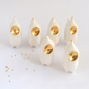 May include: Six white ceramic figurines with gold accents. Each figurine has a round, gold-colored face and a pointed top. The figurines are arranged in a semi-circle on a white surface.