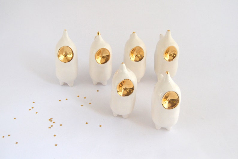 May include: Six white ceramic figurines with gold accents. Each figurine has a round, gold-colored face and a pointed top. The figurines are arranged in a semi-circle on a white surface.