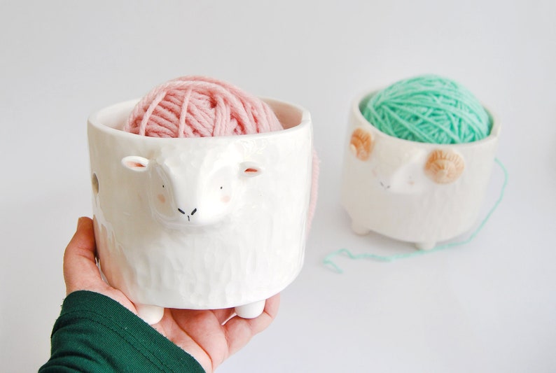 May include: Two white ceramic yarn bowls shaped like sheep, one with pink yarn and the other with green yarn.