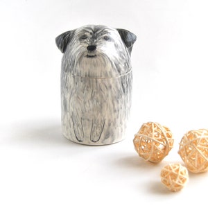 May include: A ceramic jar shaped like a small, gray and white dog with black eyes. The jar is sitting on a white surface with three small, woven, beige balls.