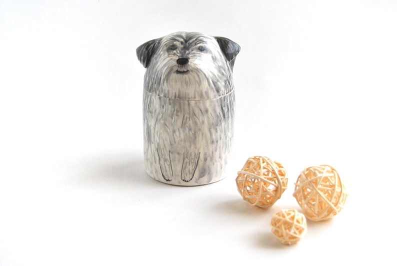 May include: A ceramic jar shaped like a small, gray and white dog with black eyes. The jar is sitting on a white surface with three small, woven, beige balls.