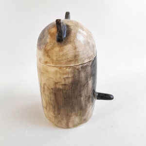 May include: A ceramic jar with a lid, painted in black and white. The jar has a rounded shape and a small handle on the side.
