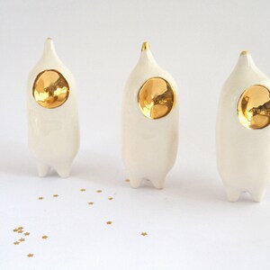 May include: Three white ceramic figurines with gold accents. Each figurine has a rounded, elongated body and a pointed head with a gold circle in the center. The figurines are arranged in a row on a white surface.