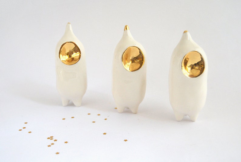 May include: Three white ceramic figurines with gold accents. Each figurine has a rounded, elongated body and a pointed head with a gold circle in the center. The figurines are arranged in a row on a white surface.