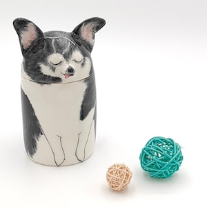 May include: A ceramic jar with a black and white dog illustration. The dog has its eyes closed and its tongue sticking out. The jar is sitting on a white surface with two small, woven balls, one teal and one beige.