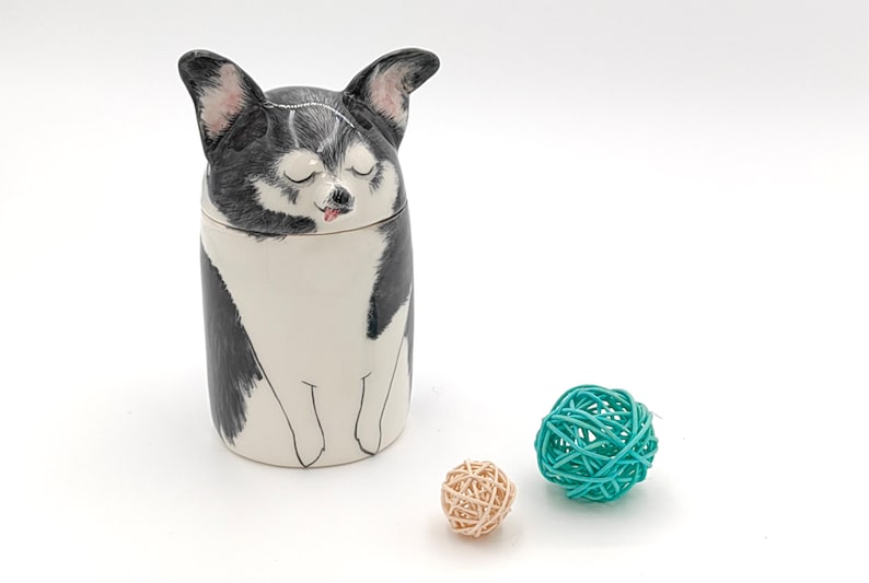 May include: A ceramic jar with a black and white dog illustration. The dog has its eyes closed and its tongue sticking out. The jar is sitting on a white surface with two small, woven balls, one teal and one beige.