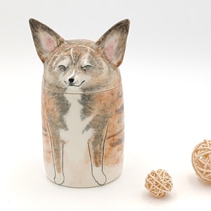 May include: A ceramic jar shaped like a dog with brown, white, and gray fur. The dog has its eyes closed and is smiling. The jar is sitting on a white surface.