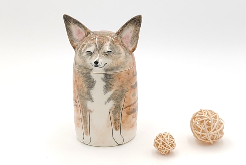 May include: A ceramic jar shaped like a dog with brown, white, and gray fur. The dog has its eyes closed and is smiling. The jar is sitting on a white surface.