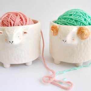 May include: Two ceramic yarn bowls shaped like sheep, one with pink yarn and the other with green yarn.