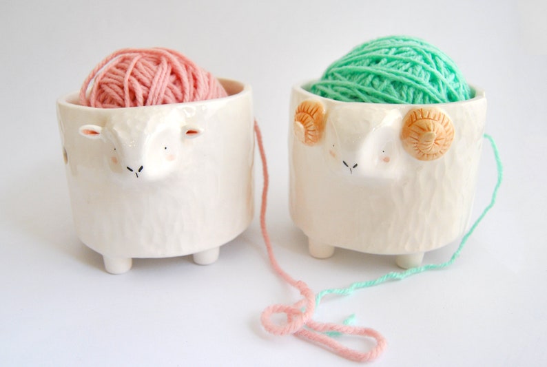 May include: Two ceramic yarn bowls shaped like sheep, one with pink yarn and the other with green yarn.