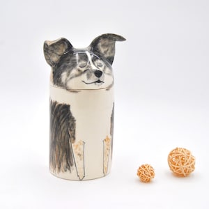 May include: Ceramic jar with a dog's face on the lid. The dog is black, white, and brown with closed eyes. The jar is white with black and brown markings. Two small, woven, light brown balls are in the foreground.