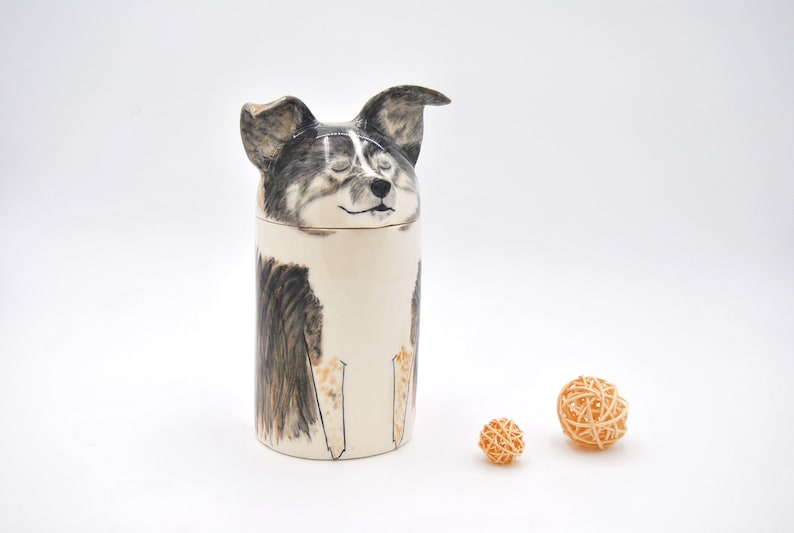 May include: Ceramic jar with a dog's face on the lid. The dog is black, white, and brown with closed eyes. The jar is white with black and brown markings. Two small, woven, light brown balls are in the foreground.