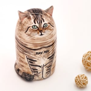 May include: A ceramic cat figurine with brown and black stripes, green eyes, and the words "May May" written on its belly. The figurine is sitting on a white surface.