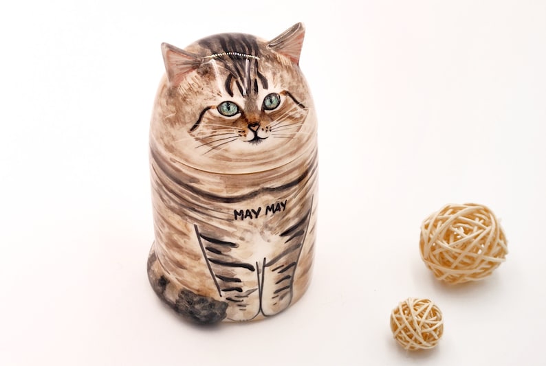 May include: A ceramic cat figurine with brown and black stripes, green eyes, and the words "May May" written on its belly. The figurine is sitting on a white surface.