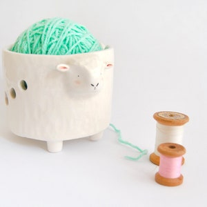 May include: White ceramic yarn bowl shaped like a sheep with a mint green ball of yarn inside. Two wooden spools of thread, one white and one pink, are on the table.