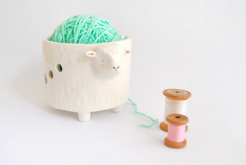 May include: White ceramic yarn bowl shaped like a sheep with a mint green ball of yarn inside. Two wooden spools of thread, one white and one pink, are on the table.