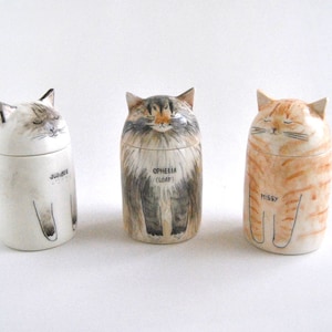 May include: Three ceramic cat figurines with painted details. The cat on the left is white with gray markings and the name "JUBILEE" written on the bottom. The cat in the middle is gray and white with the name "OPHELIA (LOAF)" written on the bottom. The cat on the right is orange and white with the name "MISSY" written on the bottom.