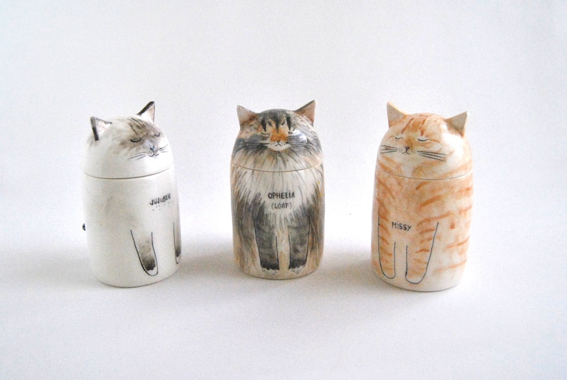 May include: Three ceramic cat figurines with painted details. The cat on the left is white with gray markings and the name "JUBILEE" written on the bottom. The cat in the middle is gray and white with the name "OPHELIA (LOAF)" written on the bottom. The cat on the right is orange and white with the name "MISSY" written on the bottom.