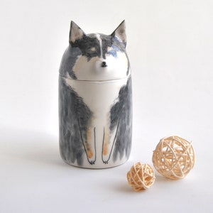 May include: Ceramic jar shaped like a dog with black, white, and brown markings. The jar has a lid and is sitting on a white surface with two small, woven balls.