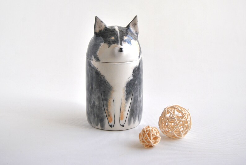 May include: Ceramic jar shaped like a dog with black, white, and brown markings. The jar has a lid and is sitting on a white surface with two small, woven balls.