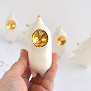 May include: A set of four white ceramic figurines with gold accents. Each figurine has a round, gold-colored face and a unique shape. The figurines are arranged on a white surface.