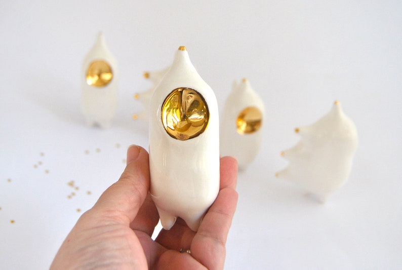May include: A set of four white ceramic figurines with gold accents. Each figurine has a round, gold-colored face and a unique shape. The figurines are arranged on a white surface.