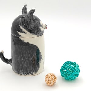 May include: A ceramic mug shaped like a black and white dog with a tail handle. The mug is sitting on a white surface with two small woven balls, one teal and one beige.