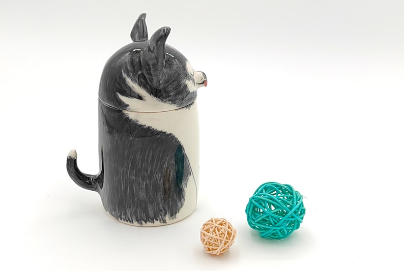 May include: A ceramic mug shaped like a black and white dog with a tail handle. The mug is sitting on a white surface with two small woven balls, one teal and one beige.