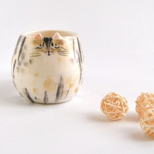 May include: A ceramic cat-shaped planter with a white and gray striped pattern and a black and white painted face. The planter is sitting on a white surface with three small, woven balls.