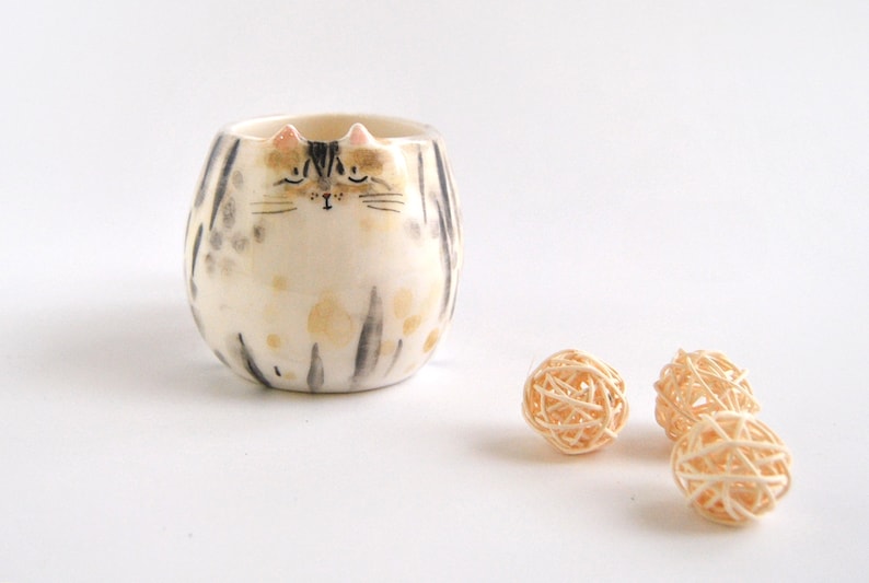 May include: A ceramic cat-shaped planter with a white and gray striped pattern and a black and white painted face. The planter is sitting on a white surface with three small, woven balls.