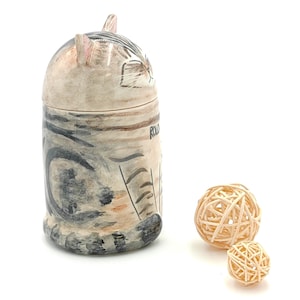 May include: A ceramic cat-shaped jar with a black and white striped pattern. The jar is sitting on a white surface with two small, woven balls in front of it.