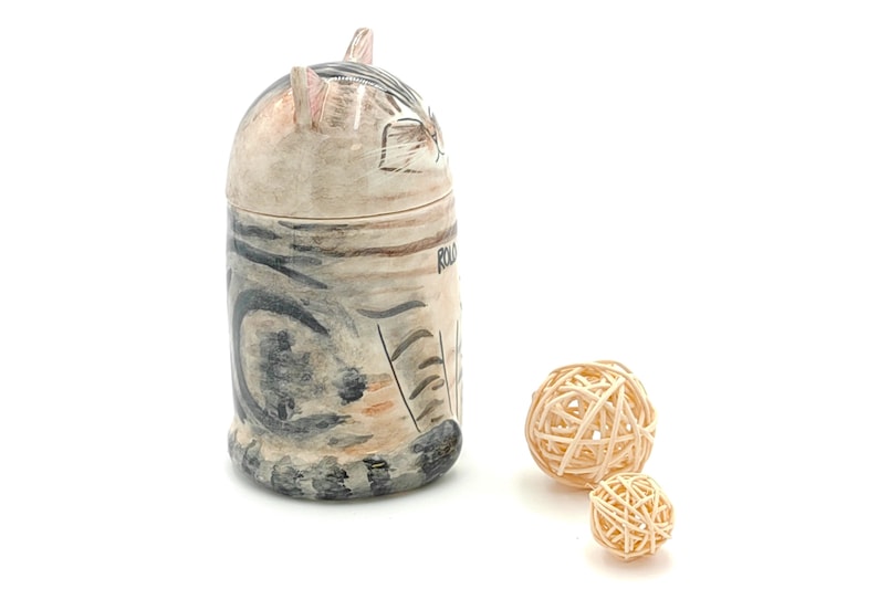 May include: A ceramic cat-shaped jar with a black and white striped pattern. The jar is sitting on a white surface with two small, woven balls in front of it.