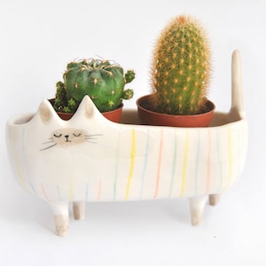 Op de afbeelding: Een witte keramische kattenplanter met pastelkleurige strepen, met twee kleine cactussen in potten. De katvormige planter heeft een staart en poten, waardoor het een uniek decoratief stuk is voor in huis.