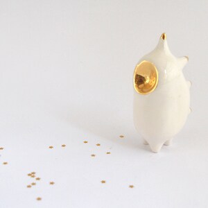 May include: White ceramic figurine with a gold accent on the head, resembling a stylized animal. The figurine is standing on a white surface with gold glitter scattered around it.