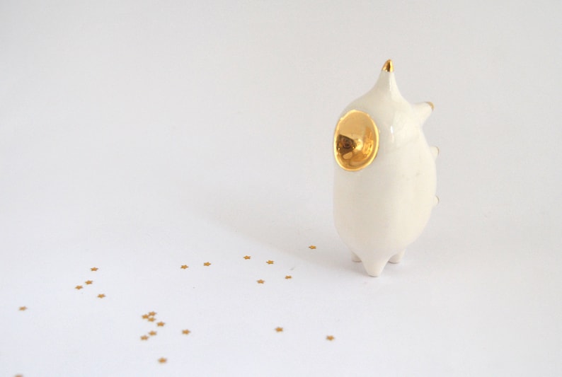 May include: White ceramic figurine with a gold accent on the head, resembling a stylized animal. The figurine is standing on a white surface with gold glitter scattered around it.