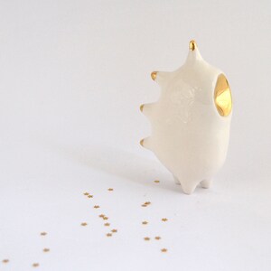 May include: A white ceramic sculpture with gold accents in the shape of a stylized animal with a single gold eye. The sculpture is sitting on a white surface with gold glitter scattered around it.