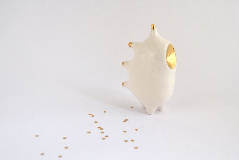 May include: A white ceramic sculpture with gold accents in the shape of a stylized animal with a single gold eye. The sculpture is sitting on a white surface with gold glitter scattered around it.