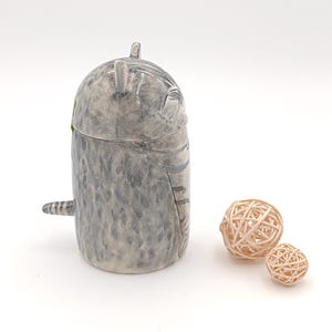 May include: A ceramic cat figurine with a gray and white speckled finish. The cat is curled up in a sleeping position with its tail wrapped around its body. The figurine is sitting on a white surface with two small, woven balls.