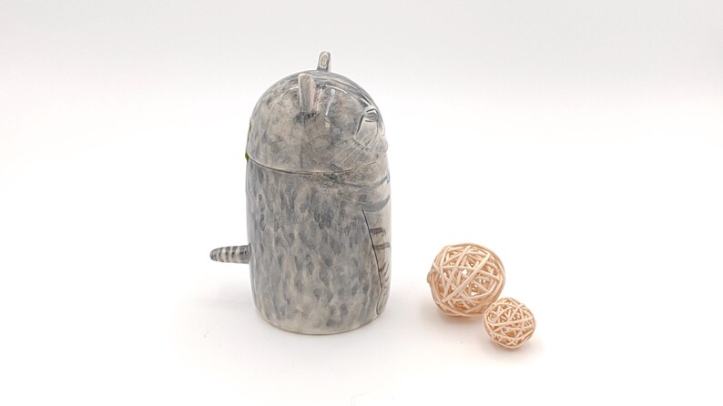 May include: A ceramic cat figurine with a gray and white speckled finish. The cat is curled up in a sleeping position with its tail wrapped around its body. The figurine is sitting on a white surface with two small, woven balls.