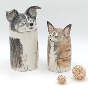 May include: Two ceramic dog figurines with painted faces. The figurines are white with black and brown markings. The figurines are sitting on a white surface with two small wooden balls.