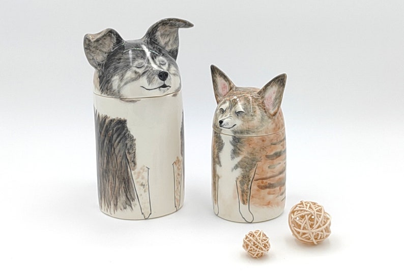 May include: Two ceramic dog figurines with painted faces. The figurines are white with black and brown markings. The figurines are sitting on a white surface with two small wooden balls.