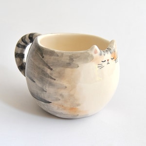 May include: A ceramic mug shaped like a sleeping cat with a gray and white striped tail for a handle. The cat has a white face with closed eyes, a black nose, and a black mouth.