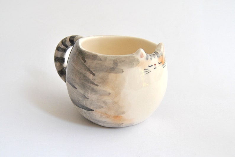 May include: A ceramic mug shaped like a sleeping cat with a gray and white striped tail for a handle. The cat has a white face with closed eyes, a black nose, and a black mouth.