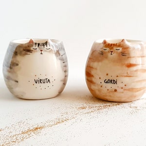 May include: Two ceramic cat mugs with painted details. The gray mug has the name "Viruta" written on it and the brown mug has the name "Gordi" written on it.